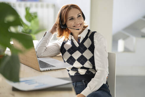 Businesswoman with headset sitting at desk with laptop in home office - JCCMF04800