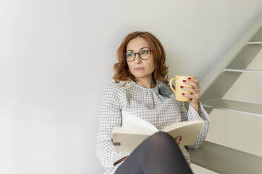 Nachdenkliche Frau mit Tagebuch und Tasse auf der Treppe zu Hause - JCCMF04824