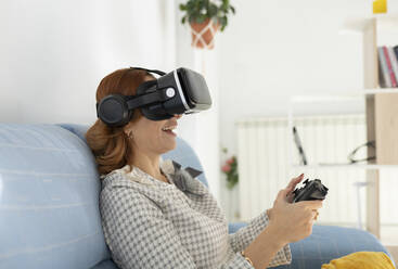Woman holding joystick wearing virtual reality headset at home - JCCMF04841