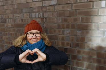 Senior woman making heart shape with hands in front of brick wall - IHF00719