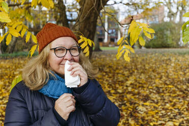 Senior woman blowing nose with tissue paper in autumn park - IHF00724