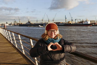Smiling senior woman making heart shape with hands at harbor - IHF00740