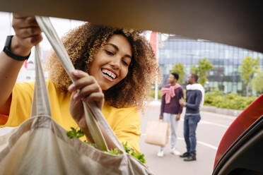 Smiling woman loading shopping bag in car trunk and friends standing in background - VYF00785