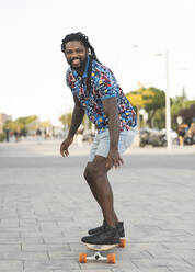 Ganzkörperansicht eines fröhlichen, sportlichen Afroamerikaners in Freizeitkleidung, der auf einem gepflasterten Gehweg auf einer Straße in der Stadt Longboard fährt - ADSF32782