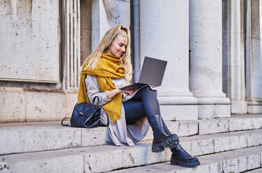Positive female freelancer in casual warm outfit checking email in laptop while sitting on stairs of building - ADSF32860