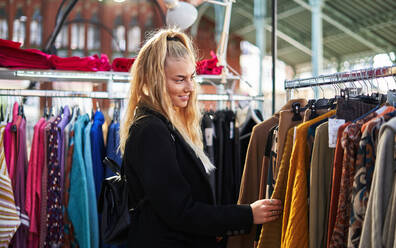 Seitenansicht einer jungen lächelnden Frau mit Rucksack, die bunte, warme Kleidung auswählt, die auf Regalen in einem Geschäft hängt - ADSF32867