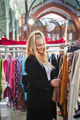 Side view of young smiling woman with backpack choosing colorful warm clothes hanging on racks in store - ADSF32868