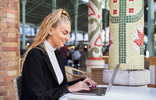 Side view of concentrated female freelancer sitting with coffee at table working on laptop in cafe - ADSF32869