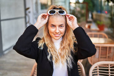 Lächelnde junge blonde Frau in eleganter schwarzer Jacke und Sonnenbrille sitzt am Tisch auf der Veranda eines Cafés - ADSF32872