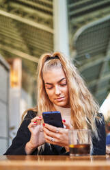 Positive blonde Frau in Freizeitkleidung, die eine Textnachricht auf einem Mobiltelefon sendet, während sie am Tisch auf der Veranda einer Cafeteria sitzt - ADSF32873