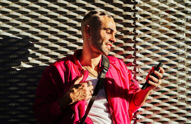 Selbstbewusster, exzentrischer Mann mit gefärbten Haaren und stilvollem Outfit steht im Sonnenlicht und surft auf seinem Handy - ADSF32881