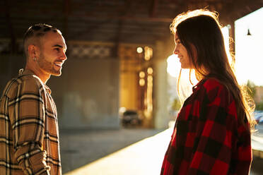 Side view of positive friends in checkered shirts looking at each other with wide smiles under bridge at sunset - ADSF32884