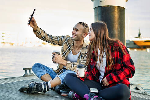 Smiling friends in roller skates sitting on quay with cups of takeaway coffee and taking self portrait on cellphone - ADSF32887