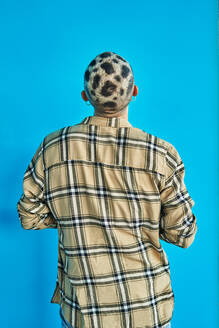 Back view of anonymous male with trendy eccentric haircut wearing checkered shirt standing against blue background - ADSF32889