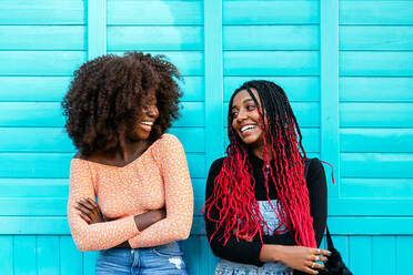 Laughing African American lady with curly hair looking at smiling black female friend against wooden blue wall looking at each other - ADSF32898