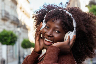 Glückliche, lockig behaarte, ethnische Dame, die Kopfhörer auf dem Kopf berührt und in die Kamera schaut, während sie auf einer verschwommenen Straße Musik hört - ADSF32905