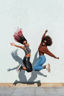 Ganzer Körper von fröhlichen afroamerikanischen Freundinnen mit Sonnenbrillen, die synchron mit ausgebreiteten Armen gegen eine weiße Wand auf der Straße springen - ADSF32906