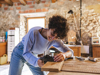 Happy Hispanic female jeweler in casual clothes using vise on wooden board during work in light workshop - ADSF32911