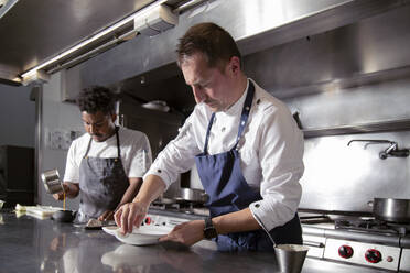 Focused professional chef wiping plate while working with black colleague pouring sauce into ceramic pot in modern kitchen - ADSF32916