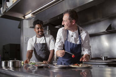 Smiling professional chef making delicious fine dish with black trainee in kitchen of restaurant and looking at each other - ADSF32920