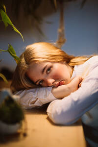 Young dissatisfied female in white blouse sitting at desk under illuminated lamp and touching chin with hand - ADSF32930