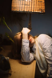 Young female in white blouse lying at desk under illuminated lamp and covering his face with his arm - ADSF32931