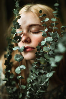 Young blond female in knitted sweater touching face with green eucalyptus twigs in hands with eyes closed - ADSF32935