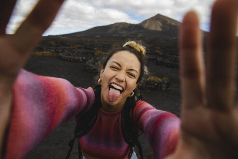 Glückliche Frau streckt die Zunge heraus und macht ein Selfie mit Landschaft im Hintergrund - MRAF00807