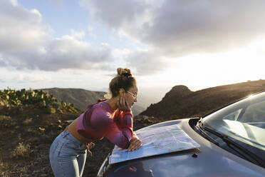 Junge Frau lehnt auf der Motorhaube eines Autos und überprüft eine Landkarte bei Sonnenuntergang - MRAF00821
