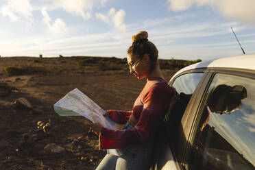 Woman with map leaning on car at sunset - MRAF00826