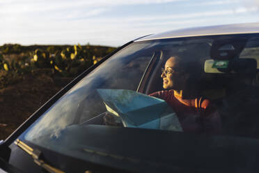 Lächelnde Frau sitzt mit Karte im Auto bei Sonnenuntergang - MRAF00828