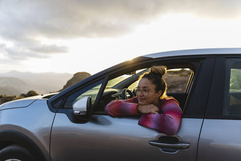 Smiling woman leaning out of car window at sunset - MRAF00831