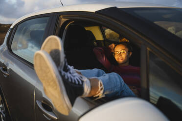 Glückliche Frau, die bei Sonnenuntergang mit hochgelegten Füßen am Autofenster sitzt - MRAF00836