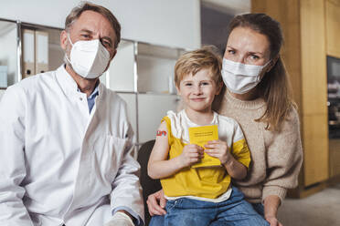 Smiling boy holding vaccination certificate with doctor and mother at center - MFF08391