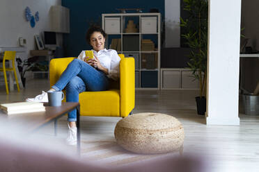 Nachdenkliche Frau sitzt mit Smartphone auf der Couch zu Hause - GIOF14591