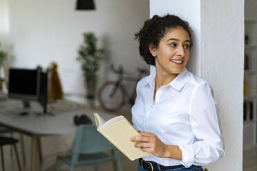 Smiling young woman leaning on column holding book at home - GIOF14596