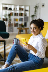 Frau mit Kaffeebecher und Smartphone zu Hause - GIOF14598