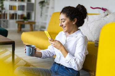 Smiling woman photographing coffee mug through smart phone at home - GIOF14599