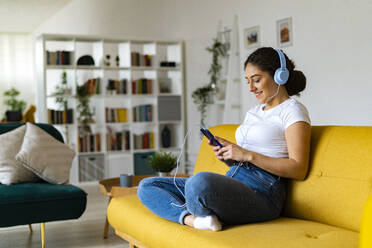 Young woman listening music through smart phone sitting on sofa at home - GIOF14603