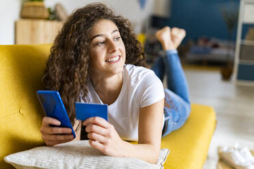 Lächelnde junge Frau mit Kreditkarte und Smartphone auf dem Sofa zu Hause liegend - GIOF14616