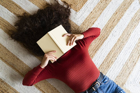 Frau, die ihr Gesicht mit einem Buch bedeckt, das zu Hause auf dem Teppich liegt - GIOF14620