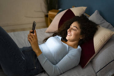 Smiling woman using smart phone lying on bed at home - GIOF14629