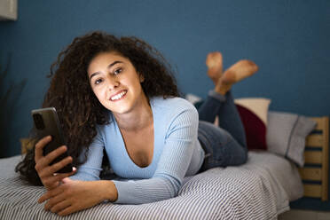 Young woman holding smart phone lying on bed - GIOF14632