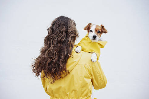 Frau mit langen braunen Haaren, die einen Hund vor einer weißen Wand trägt - EBBF05146