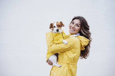 Lächelnde junge Frau, die ihren Hund an einer weißen Wand umarmt - EBBF05147