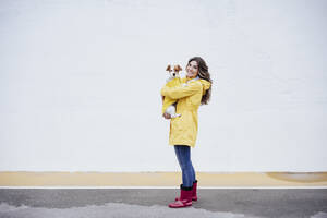 Lächelnde junge Frau im Regenmantel mit Hund auf der Straße - EBBF05148