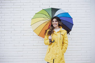 Lächelnde Frau hält mehrfarbigen Regenschirm vor einer Backsteinmauer - EBBF05149