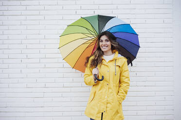 Smiling woman holding multi colored umbrella in front of brick wall - EBBF05149