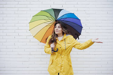 Young woman with multi colored umbrella gesturing in front of wall - EBBF05150