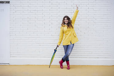 Happy woman holding umbrella on footpath in front of white brick wall - EBBF05156